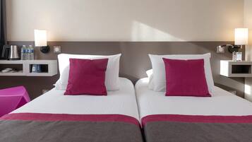 A hotel room with two beds, each with white linens and a pink pillow. There are two bedside tables with lamps, a small shelf with toiletries, and a pink chair.