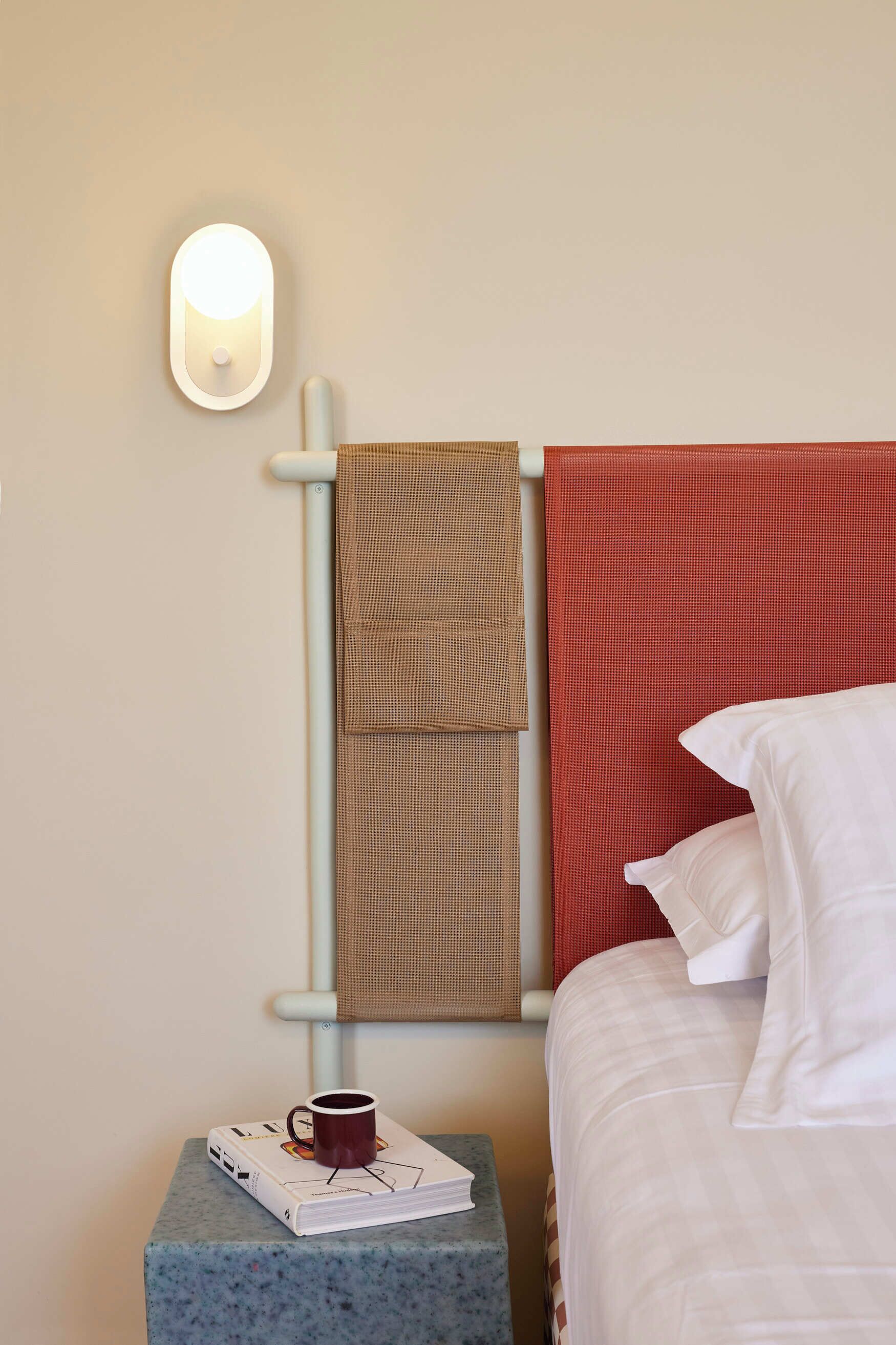 A neatly made bed with white linens, a red headboard, and a brown towel hanging on a light-colored wall. A small table beside the bed holds a book and a cup.