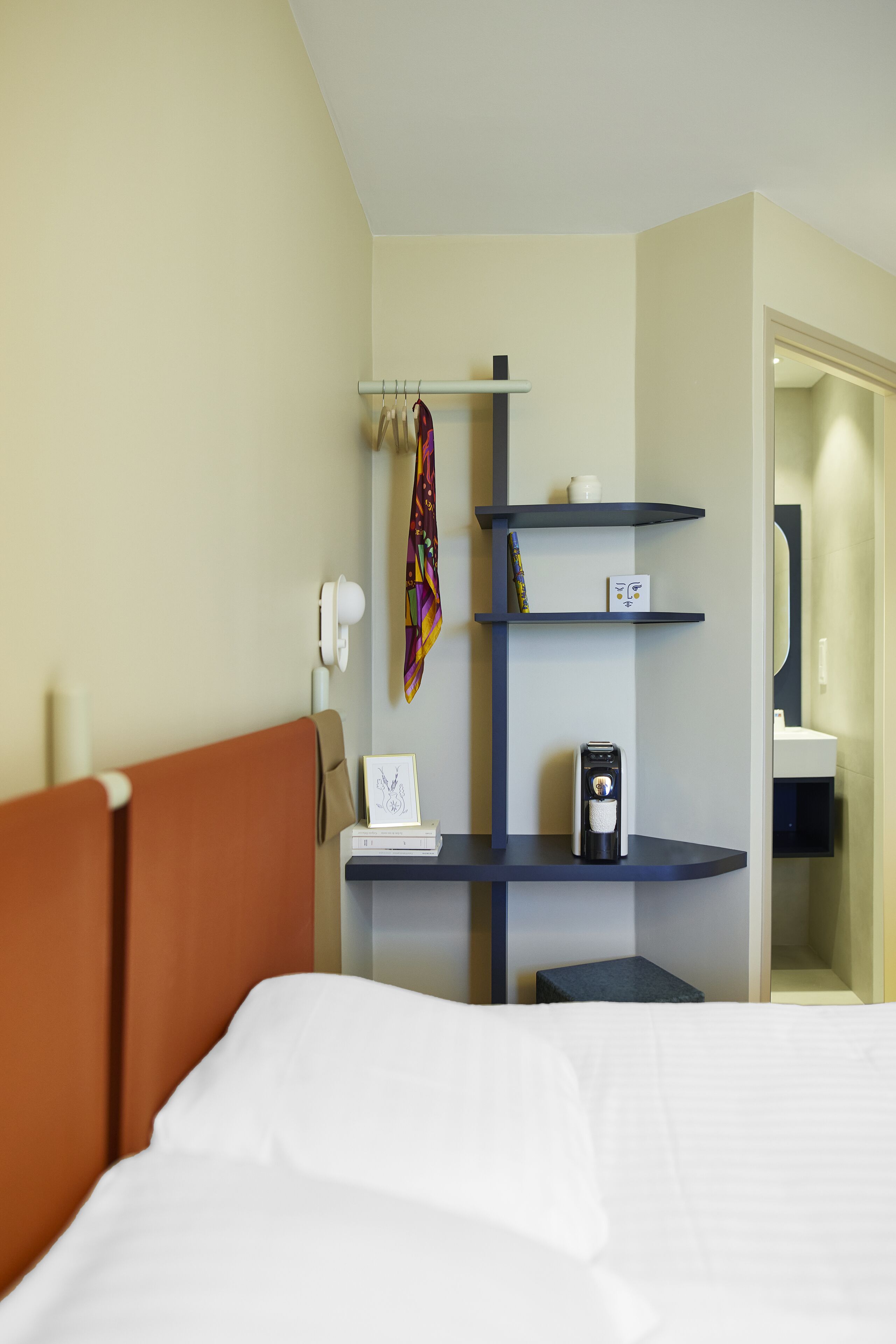 A hotel room with a bed, a desk with a lamp, a shelf with a coffee maker, and a door leading to a bathroom.