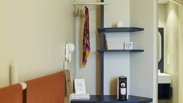 A hotel room with a bed, a desk with a lamp, a shelf with a coffee maker, and a door leading to a bathroom.