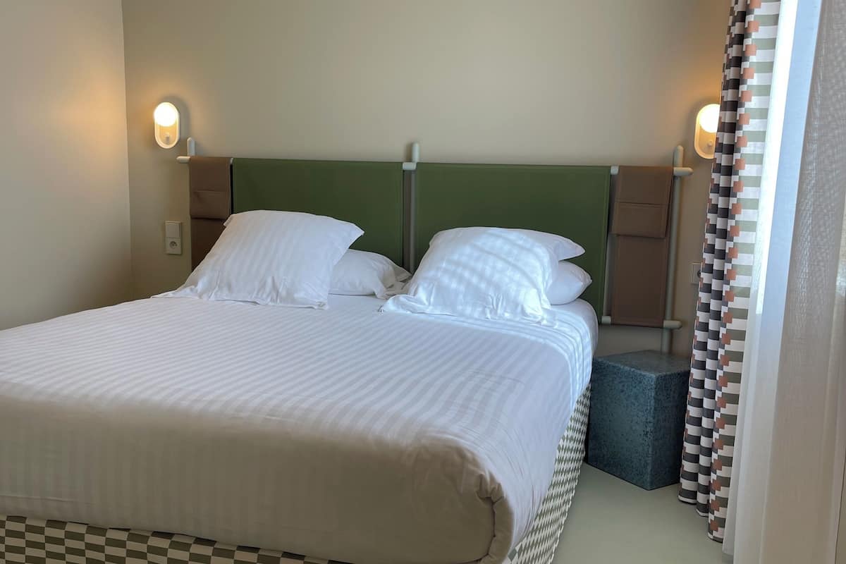 Cama bem arrumada com lençóis brancos, cabeceira verde e duas luminárias laterais.