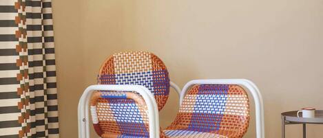 Una habitación con dos sillas tejidas, una pequeña mesa redonda con un libro y un cuadro abstracto enmarcado en la pared.