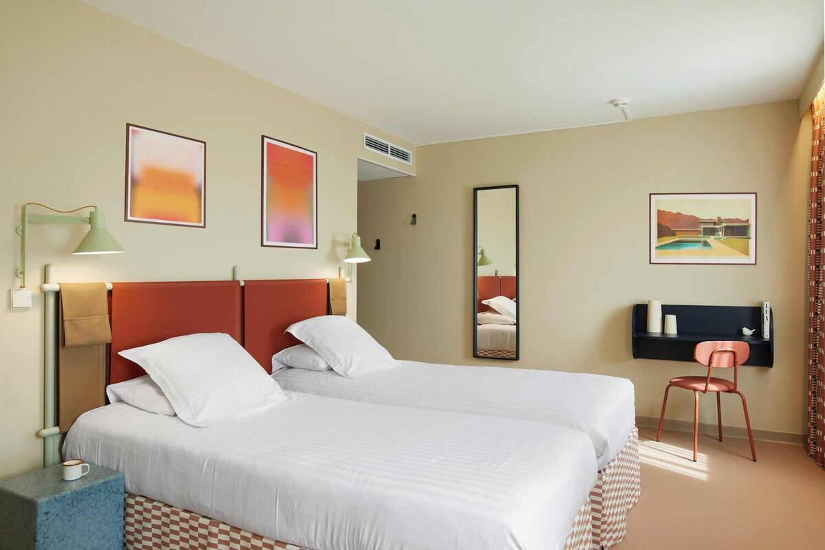 Quarto de hotel com duas camas, uma escrivaninha, uma cadeira, um espelho e obras de arte nas paredes.
