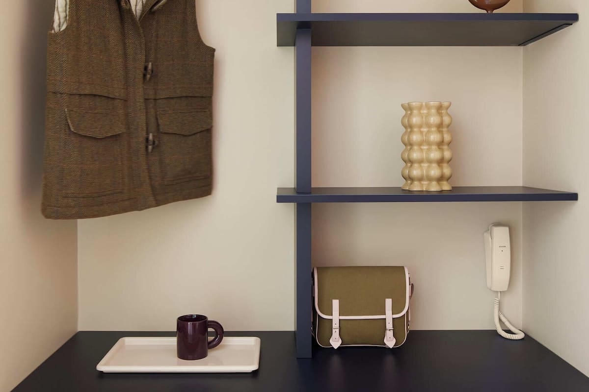 Quarto com uma escrivaninha azul escura, um cabide, um vaso, um telefone e uma caneca.