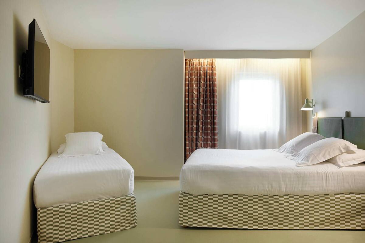 Quarto de hotel com duas camas de solteiro, uma televisão de tela plana fixada na parede e uma janela com cortinas.