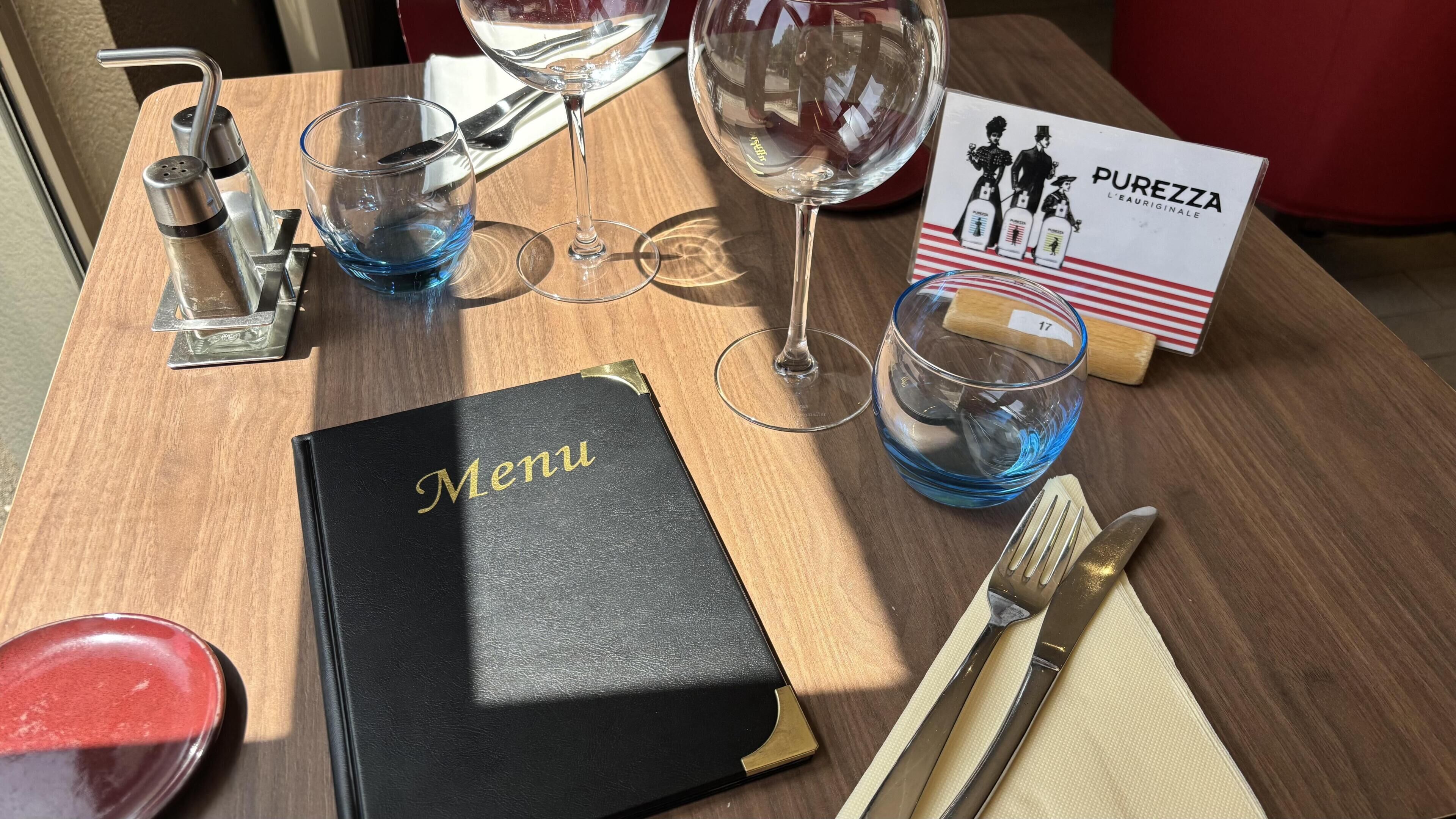 Une table en bois, sur laquelle se trouvent un présentoir à menus noir, des verres à vin, un verre à eau, une carte des menus et une carte publicitaire pour une boisson.