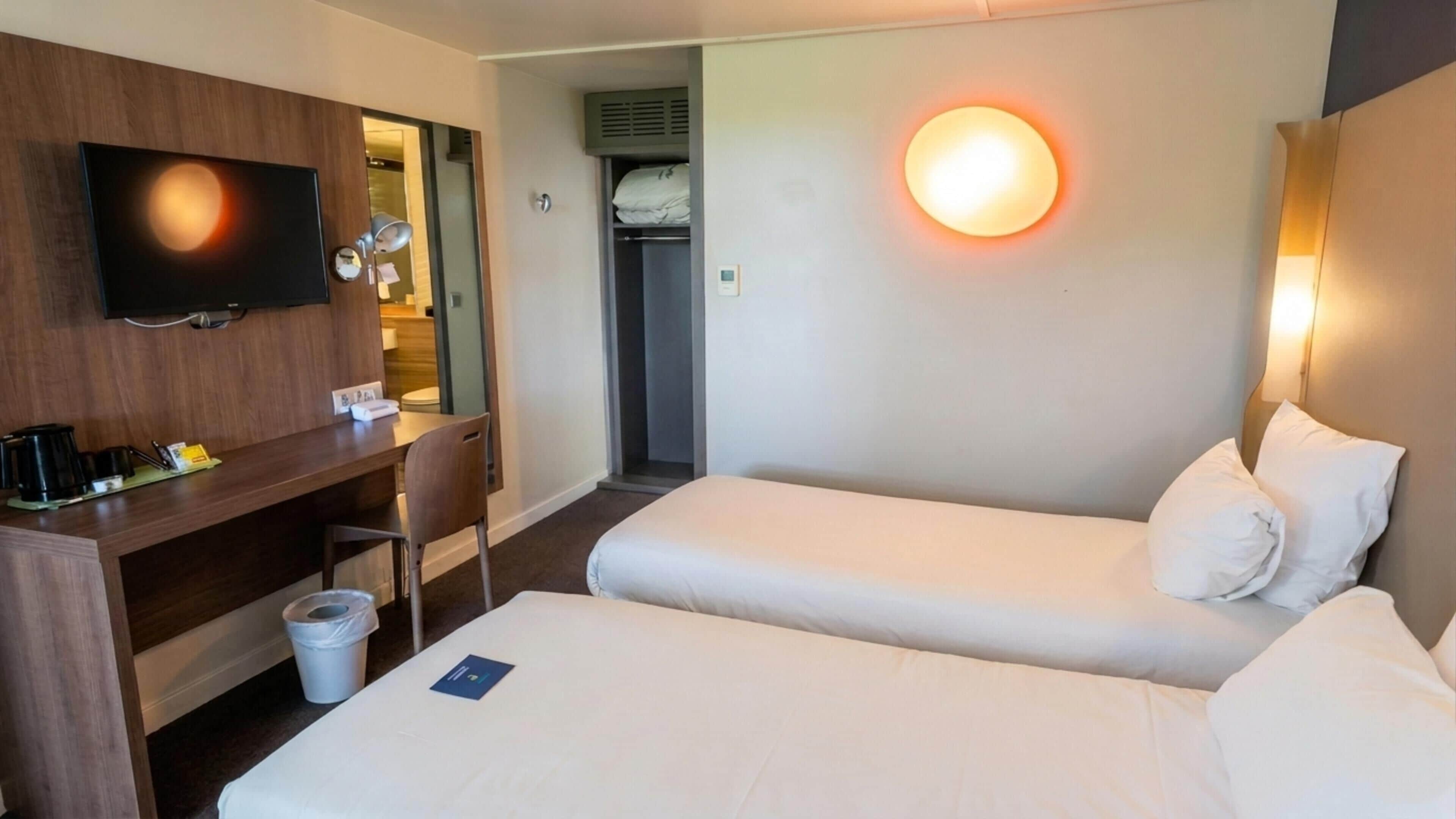 Une chambre d’hôtel équipée d’une télévision, d’un bureau, de deux lits, d’une poubelle et d’une lampe fixée au mur.