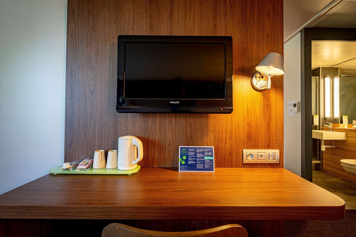 Una habitación de hotel con un escritorio de madera, un televisor de pantalla plana montado en la pared, una lámpara de noche y una mesita con una cafetera y un cartel de bienvenida.
