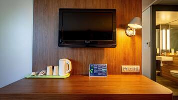 Quarto de hotel com secretária de madeira, televisão de tela plana na parede, abajur e uma pequena mesa com cafeteira e placa de boas-vindas.