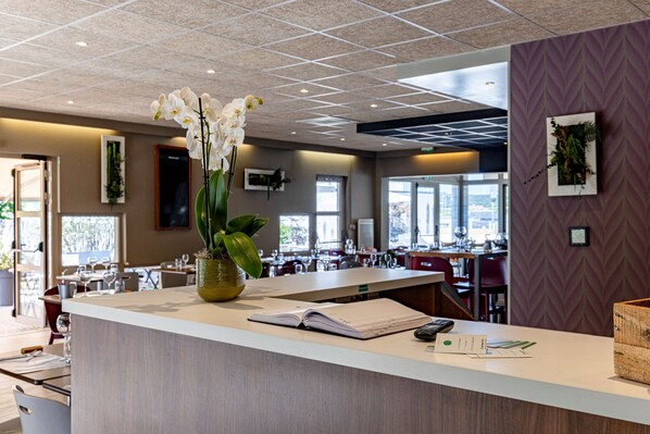 Zona de recepción de un hotel con mostrador, un jarrón de orquídeas blancas y vista al comedor.