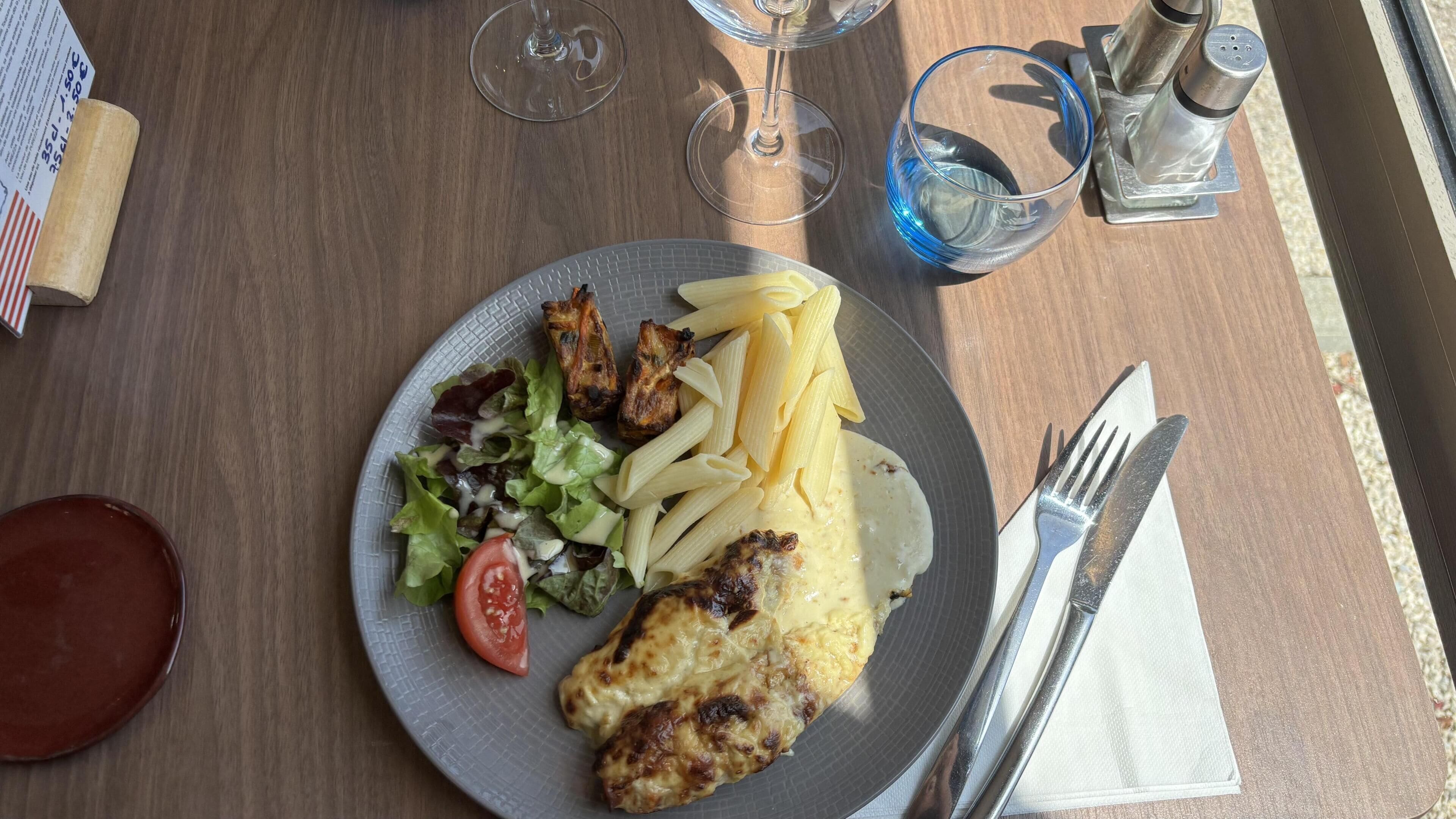 Un plato con pollo a la plancha, pasta y ensalada sobre una mesa de madera.