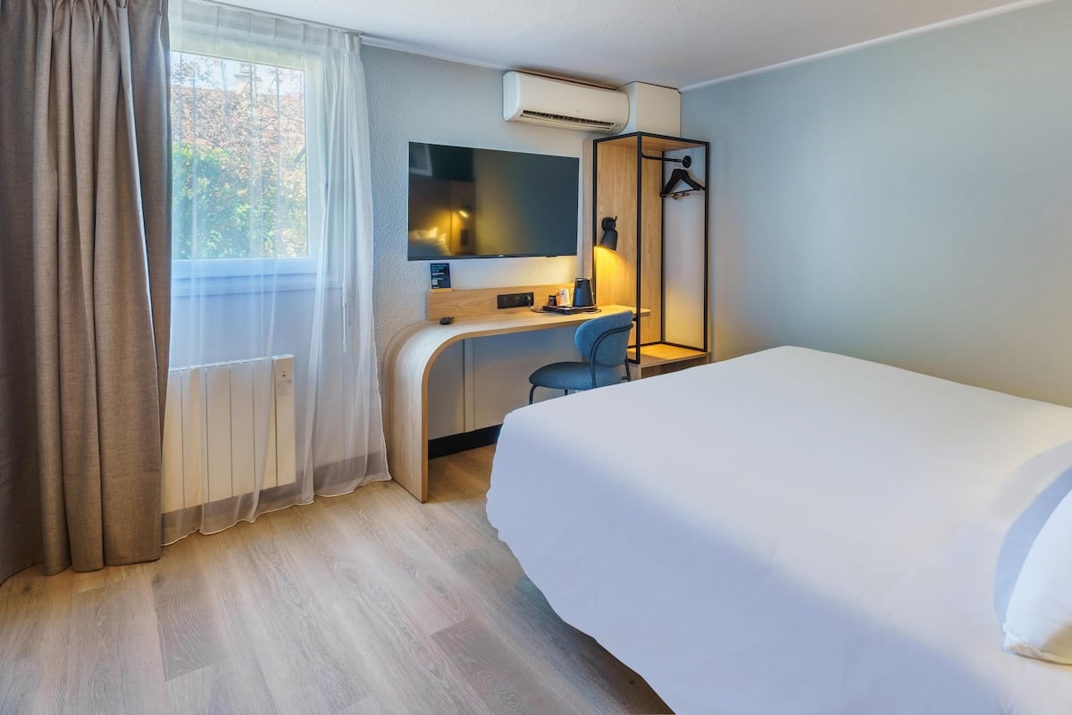 A hotel room with a bed, a desk, a chair, a mirror, a hanging light, and a window with curtains.