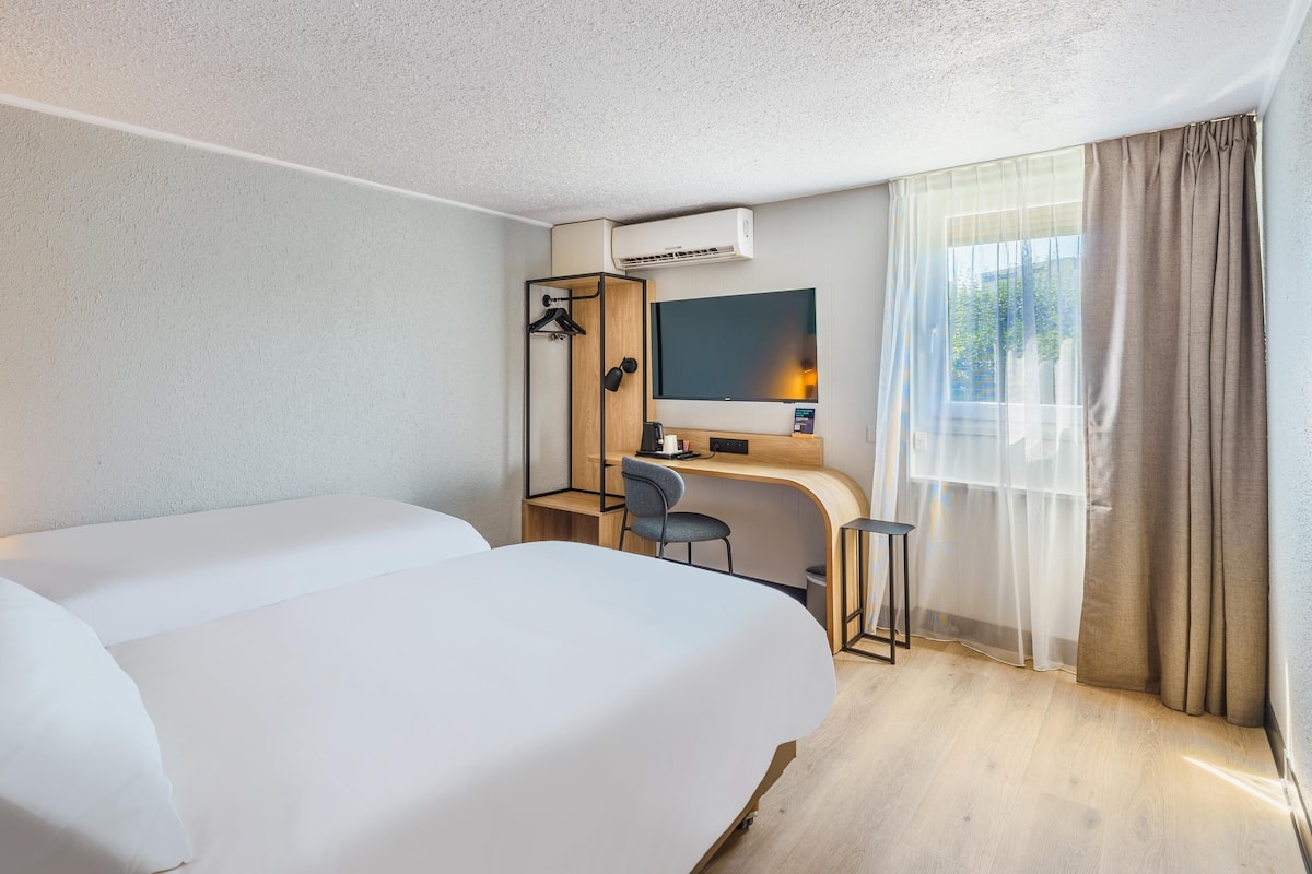 A hotel room with two beds, a desk, a chair, a TV, and an air conditioning unit.