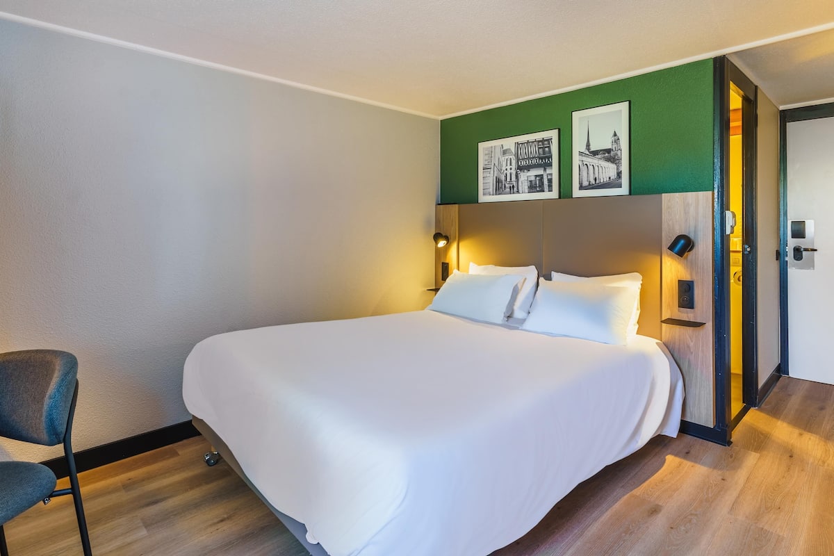 A hotel room with a large bed, a chair, a wooden floor, and a green accent wall with framed pictures.