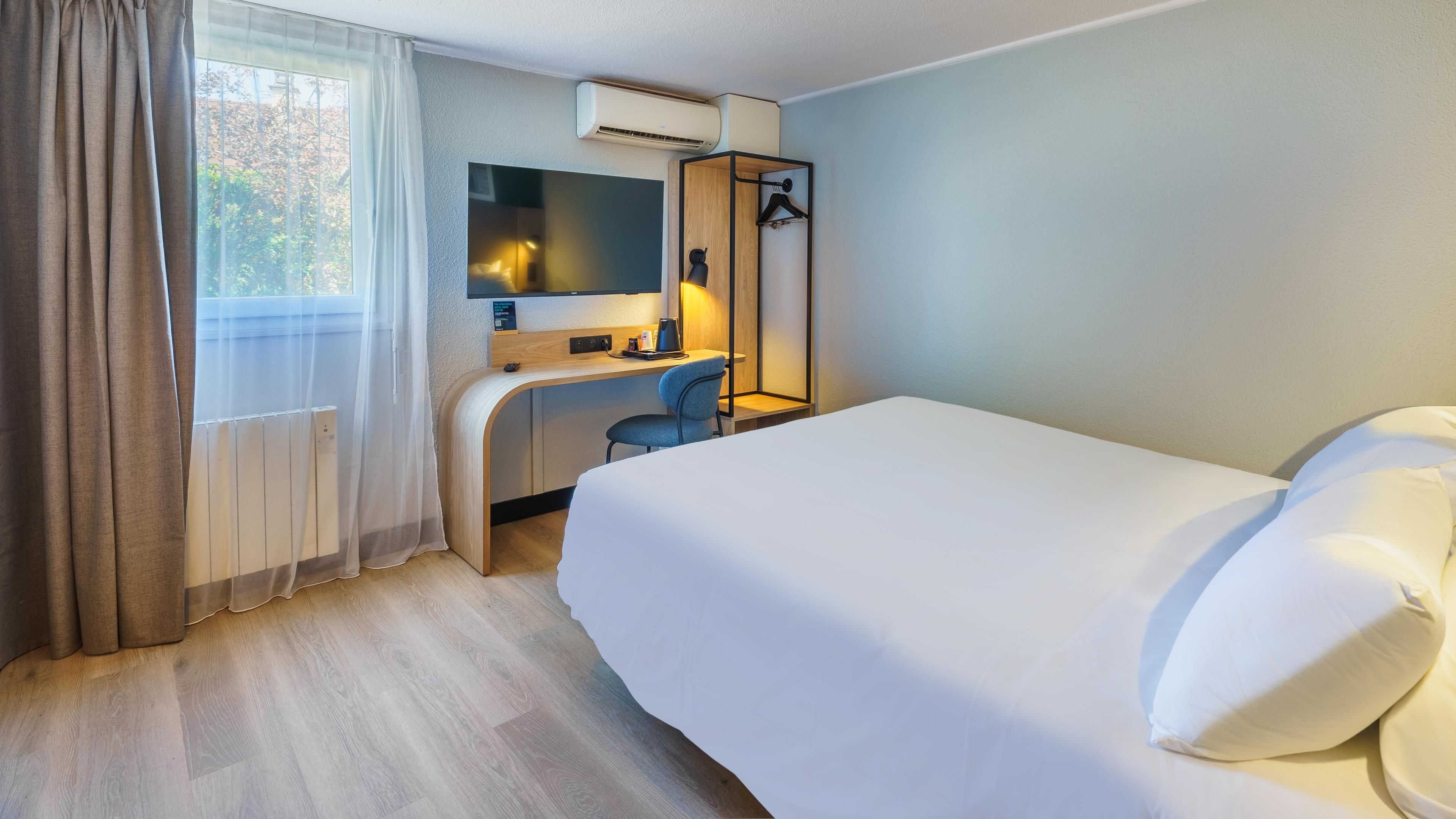 A hotel room with a bed, a desk, a chair, a mirror, a hanging light, and a window with curtains.