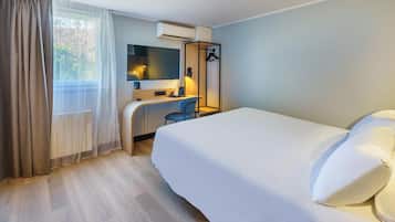 A hotel room with a bed, a desk, a chair, a mirror, a hanging light, and a window with curtains.