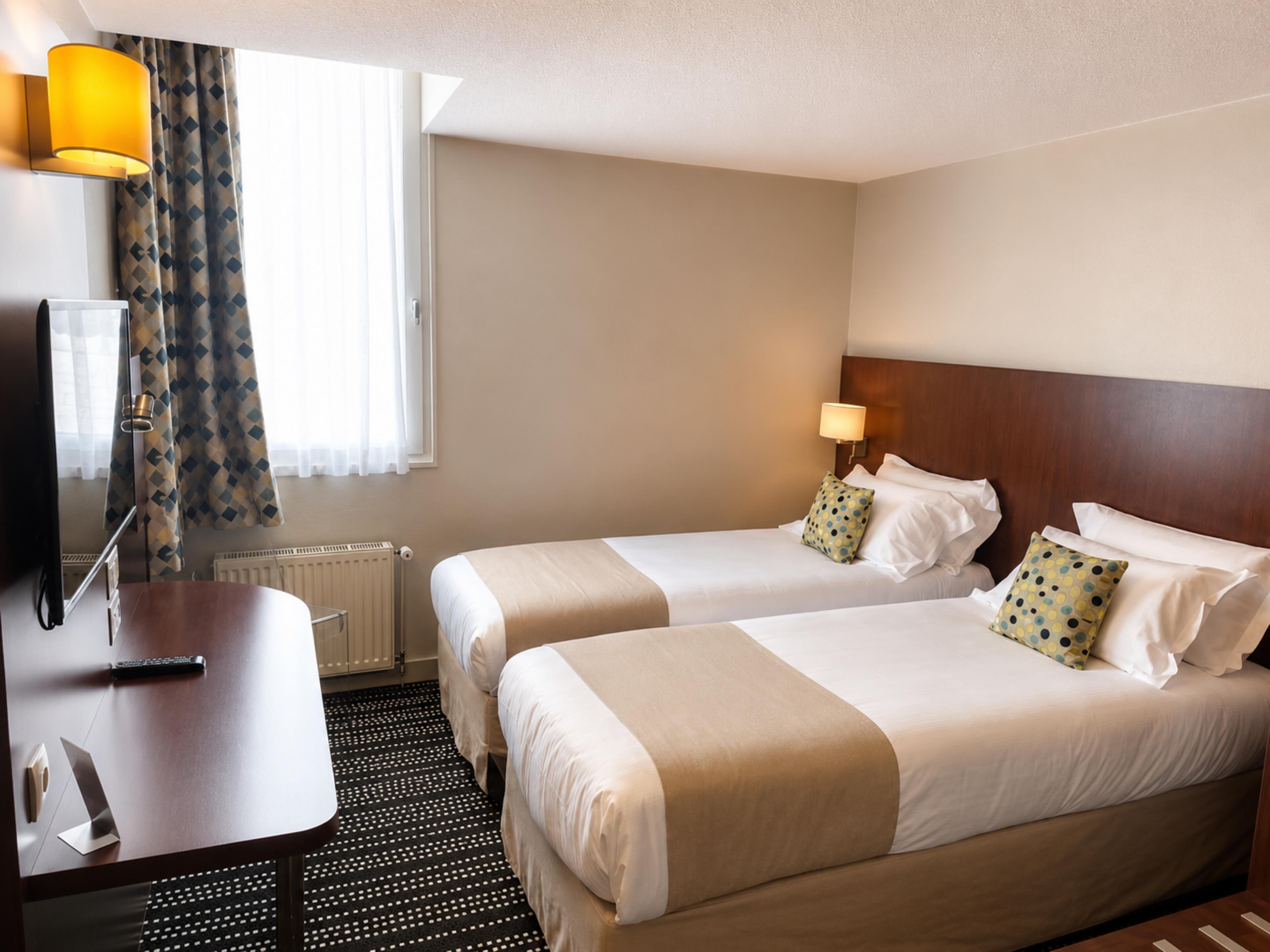 Habitación de hotel con dos camas, un escritorio, televisor y ventana con cortinas.
