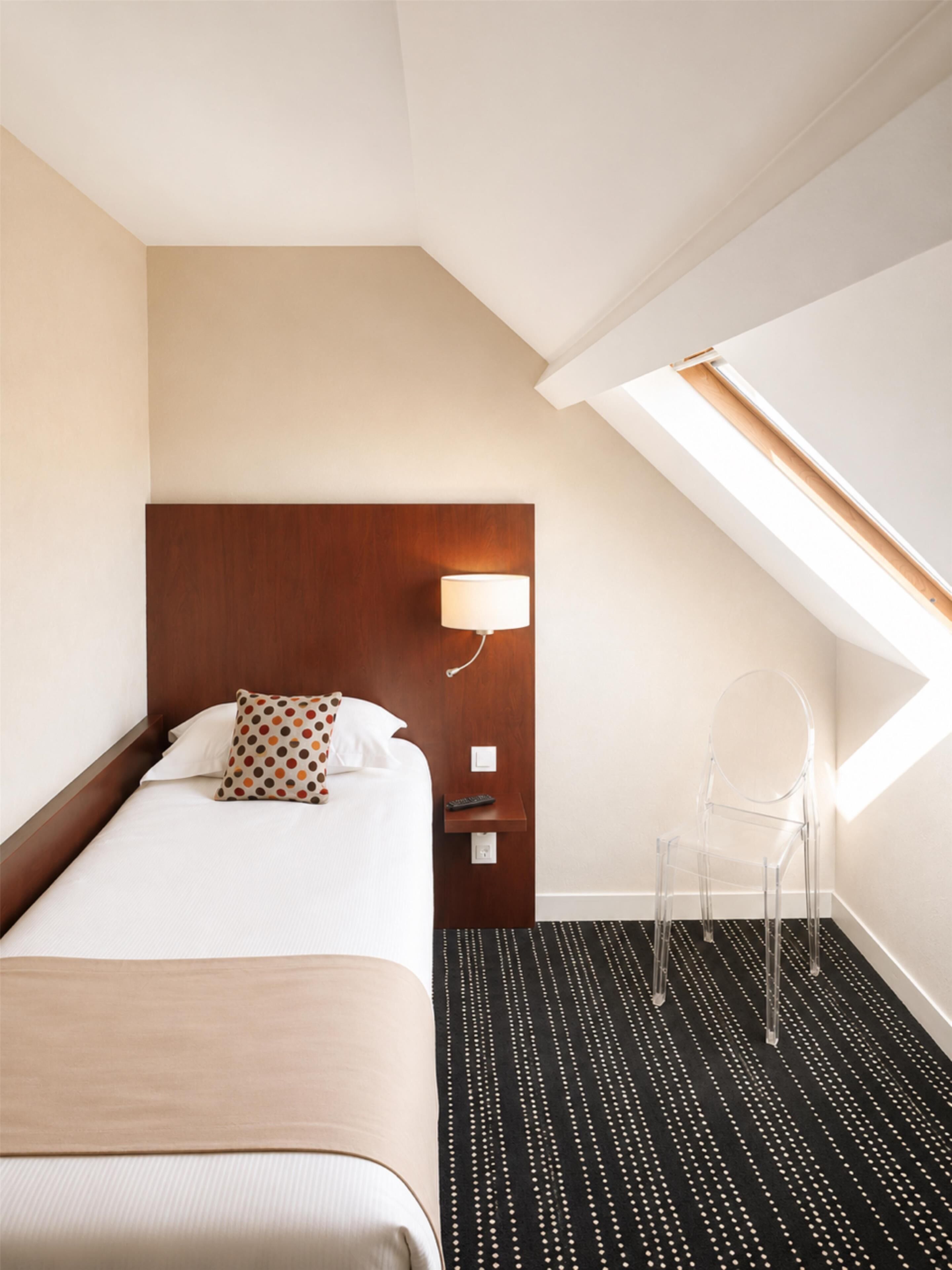Una habitación de hotel con cama, mesita de noche, lámpara, silla y tragaluz.