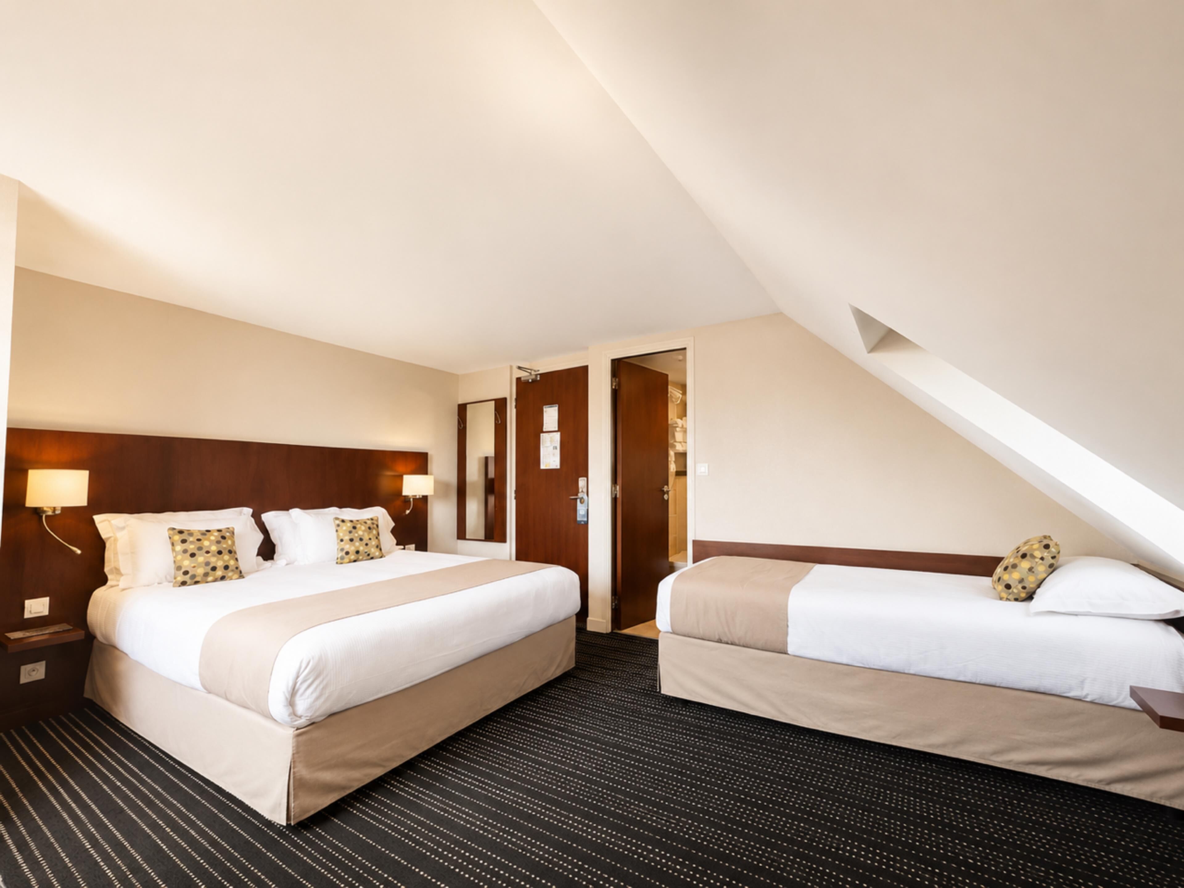 A hotel room with two beds, a dark patterned carpet, and a sloped ceiling.