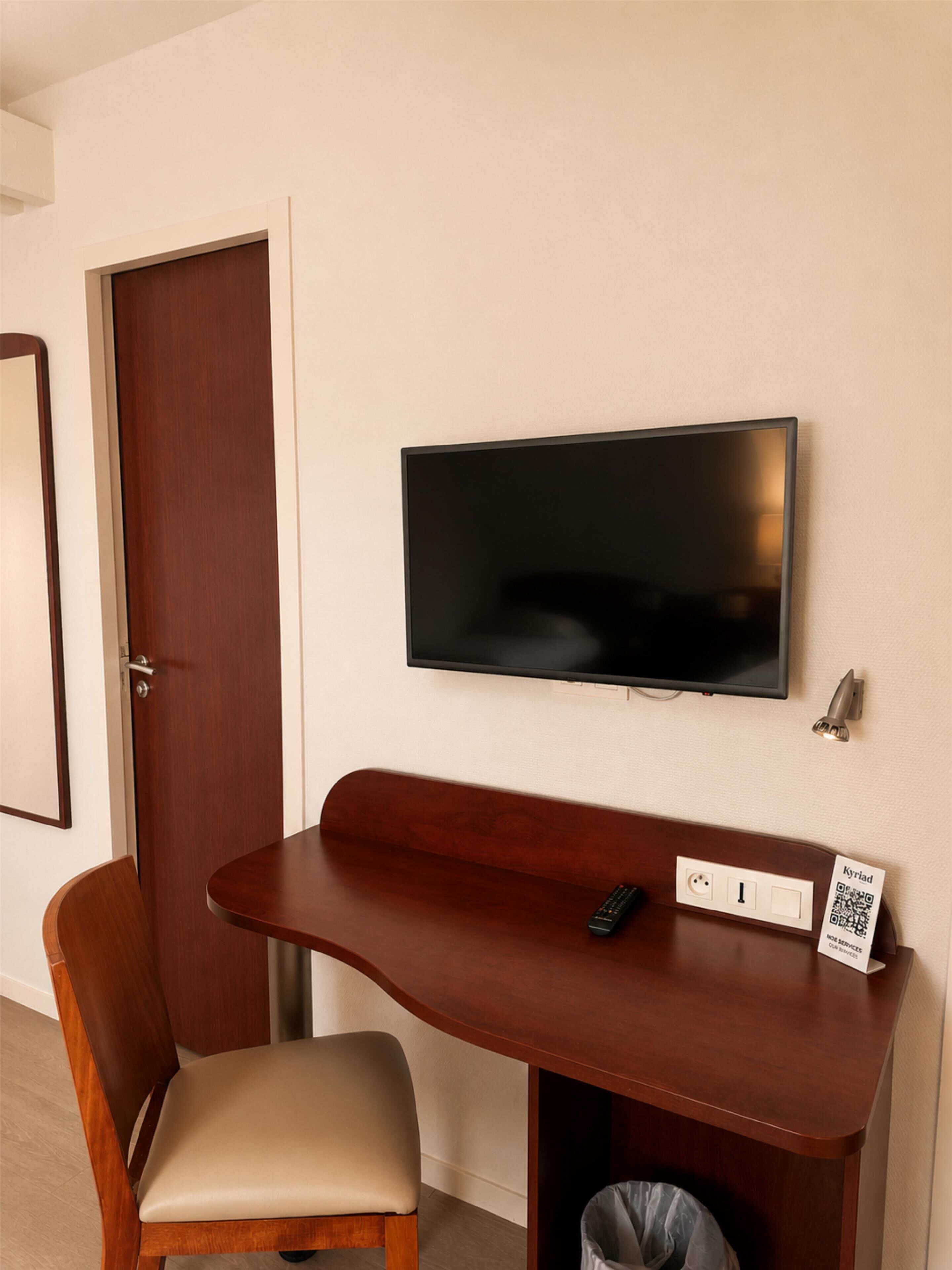 A room with a wooden desk, a chair, a television mounted on the wall, a door, and a trash bin.