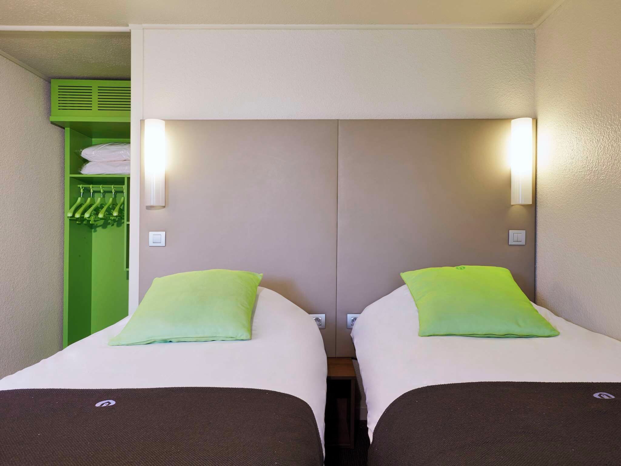 Une chambre d’hôtel avec deux lits, chacun doté d’un oreiller et d’une tête de lit. On y trouve un meuble de rangement vert où sont suspendus des vêtements, ainsi qu’un interrupteur sur le mur.
