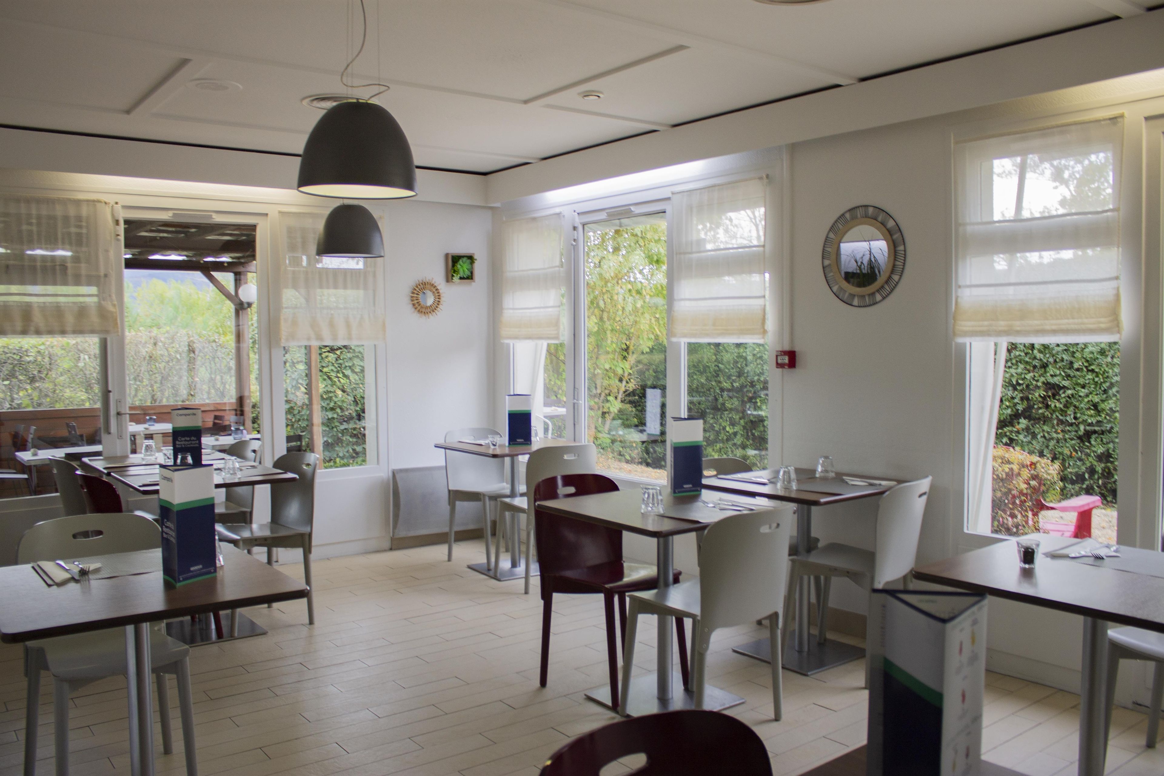 A dining area with tables and chairs, large windows with blinds, and a ceiling light fixture.