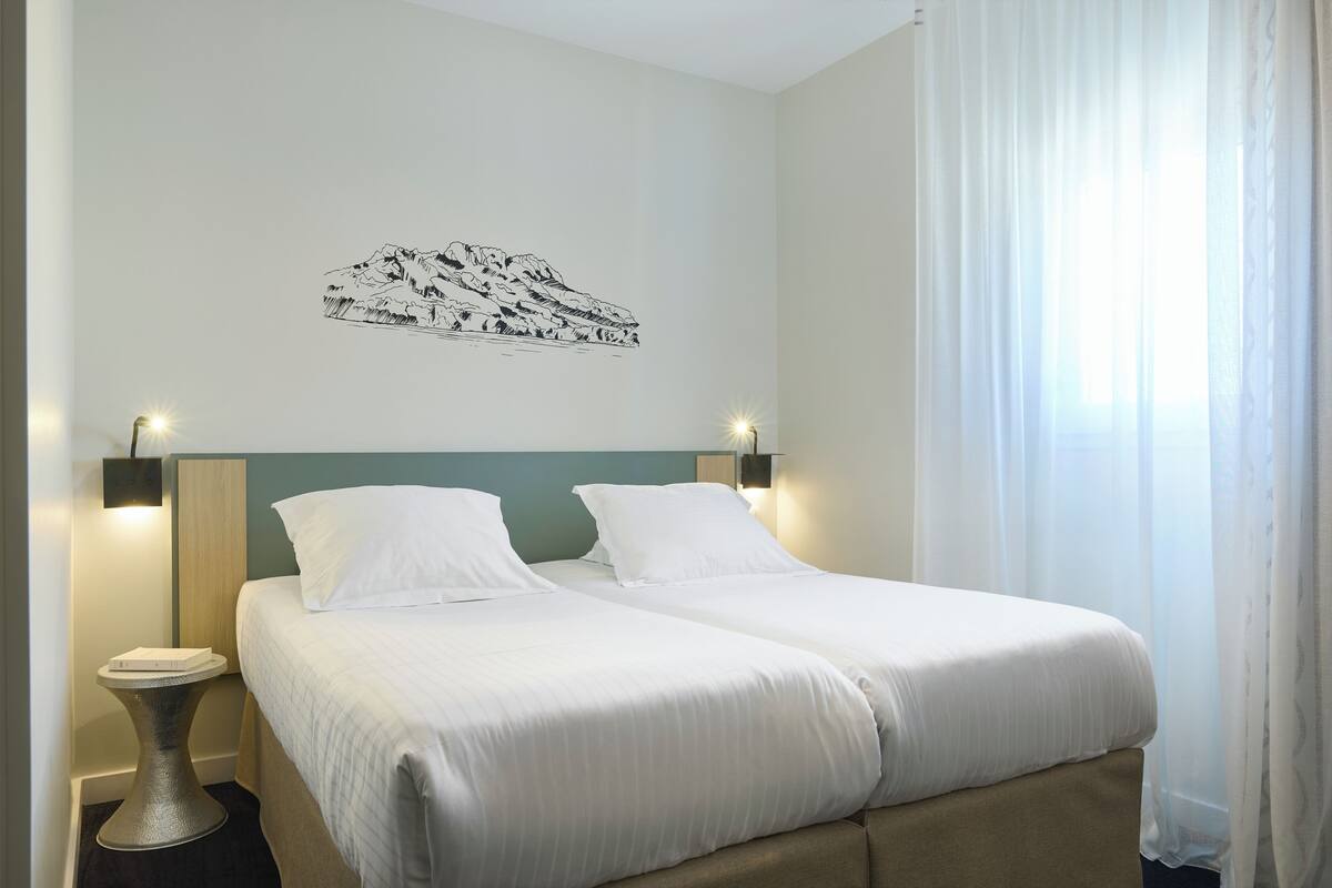 A double bed with white linens, two bedside tables with lamps, a wall-mounted artwork of a mountain scene, and a window with sheer curtains.