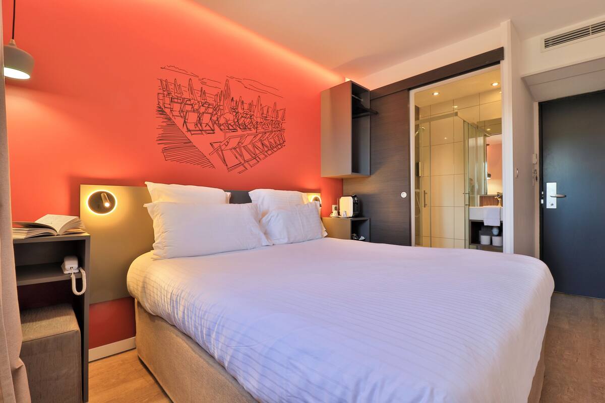 A hotel room with a large bed, a bedside table with a phone, a wall-mounted light, and a door leading to a bathroom.