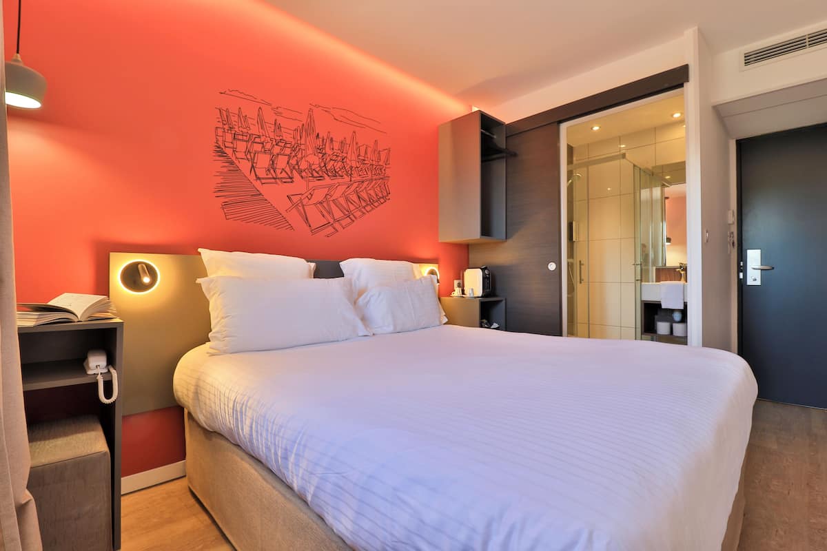 A hotel room with a large bed, a bedside table with a phone, a wall-mounted light, and a door leading to a bathroom.