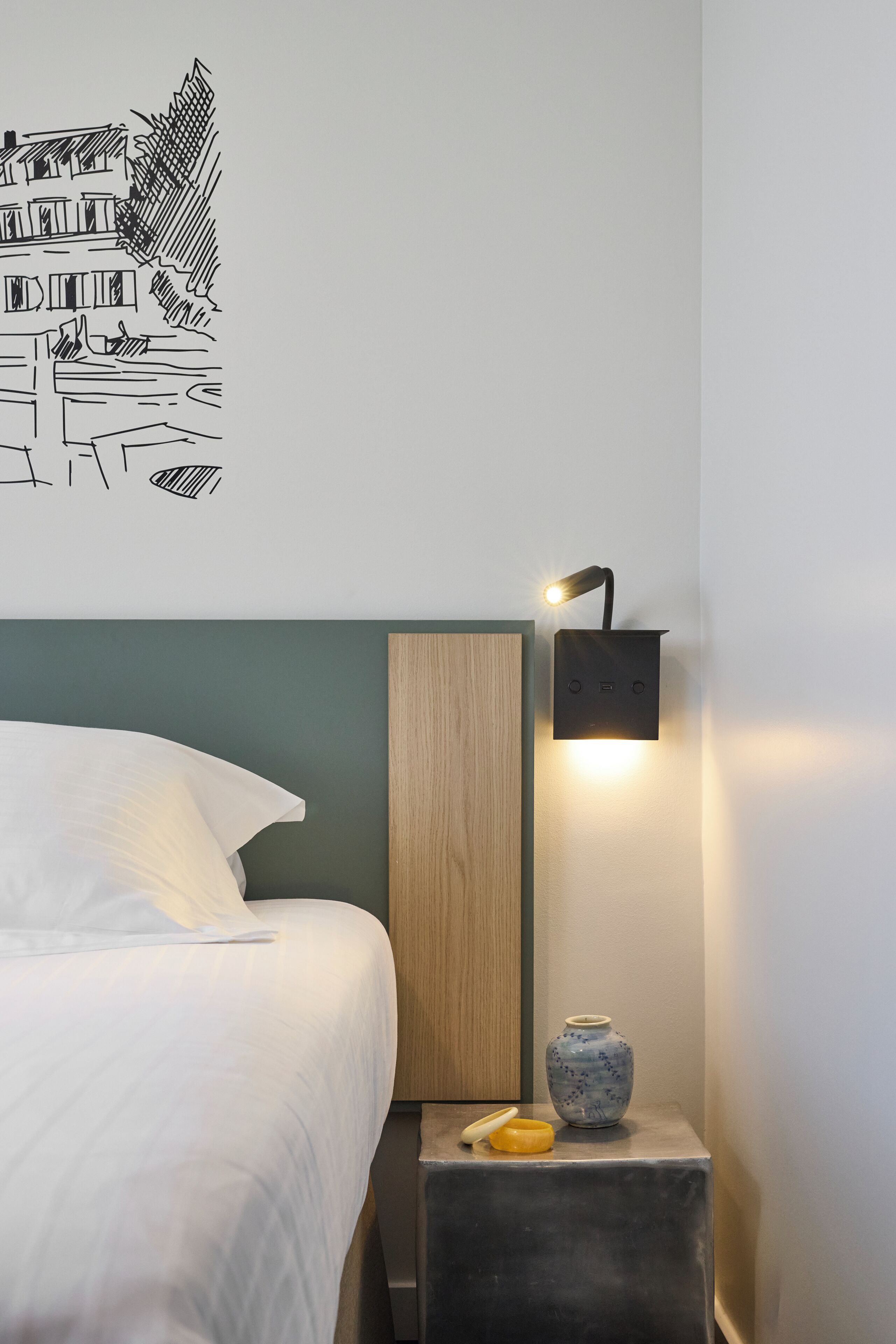 A bedroom with a bed, a wall-mounted light, a small table with a vase, and a wall art piece.
