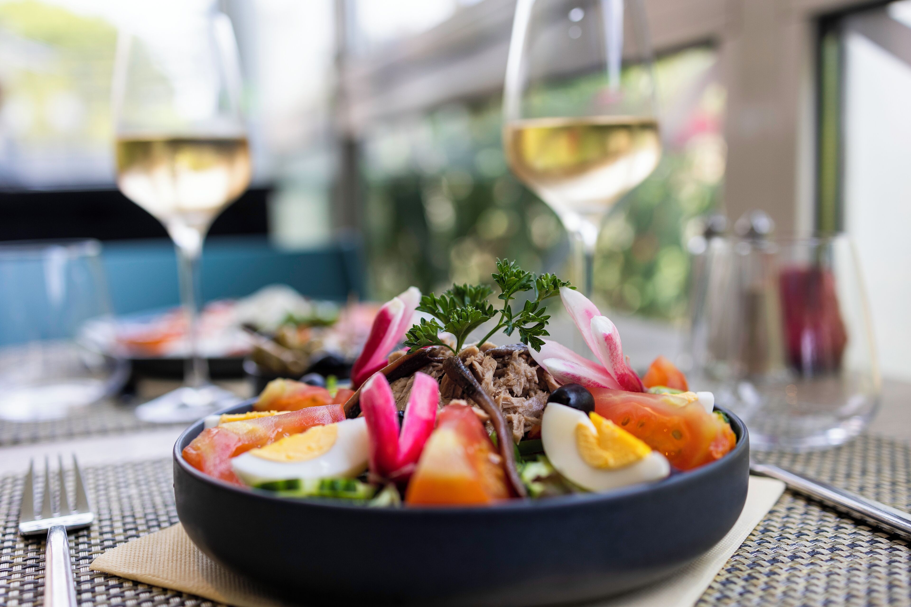 Un saladier rempli de salade avec divers ingrédients, deux verres de vin blanc et une table dressée.