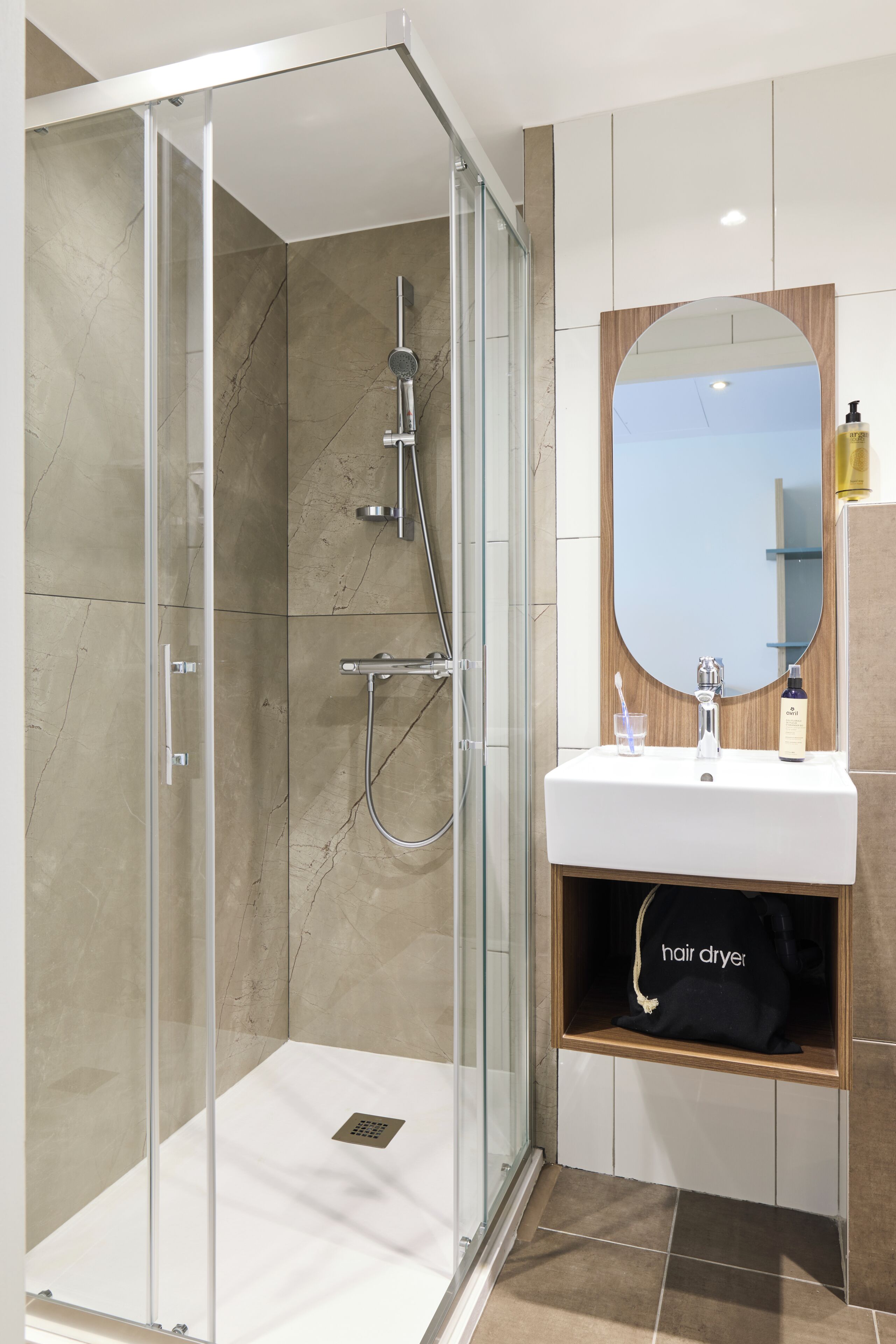 Une salle de bain moderne avec une douche à parois de verre, un lavabo avec un miroir et un sèche-cheveux posé sur l’étagère.