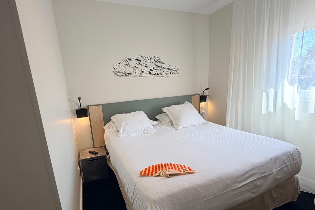 A hotel room with a bed, white curtains, a wall-mounted lamp, and a mountain landscape artwork.