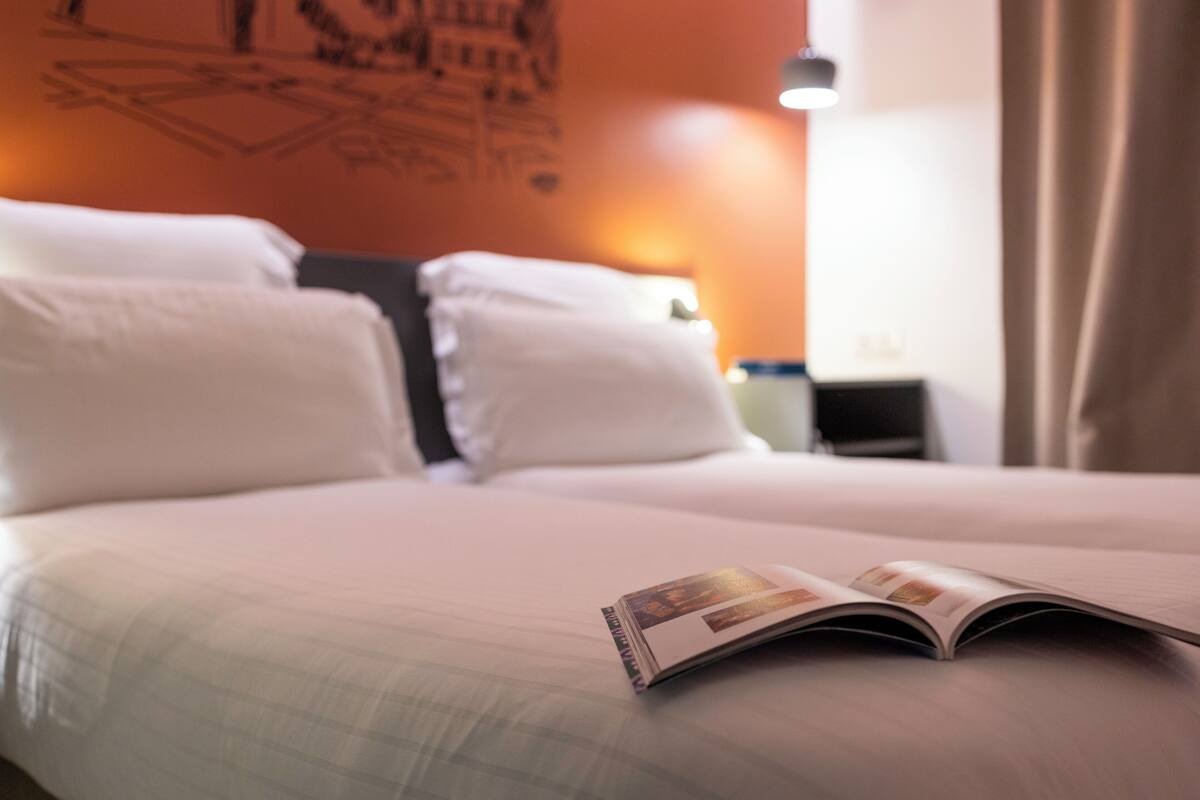 A neatly made bed with a magazine on it, a nightstand with a lamp, and a wall with a geometric design.