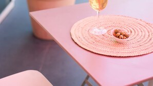 A table with a glass of drink and a small bowl of nuts, a potted plant, and a pink tablecloth on a balcony.
