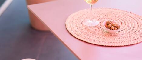A table with a glass of drink and a small bowl of nuts, a potted plant, and a pink tablecloth on a balcony.
