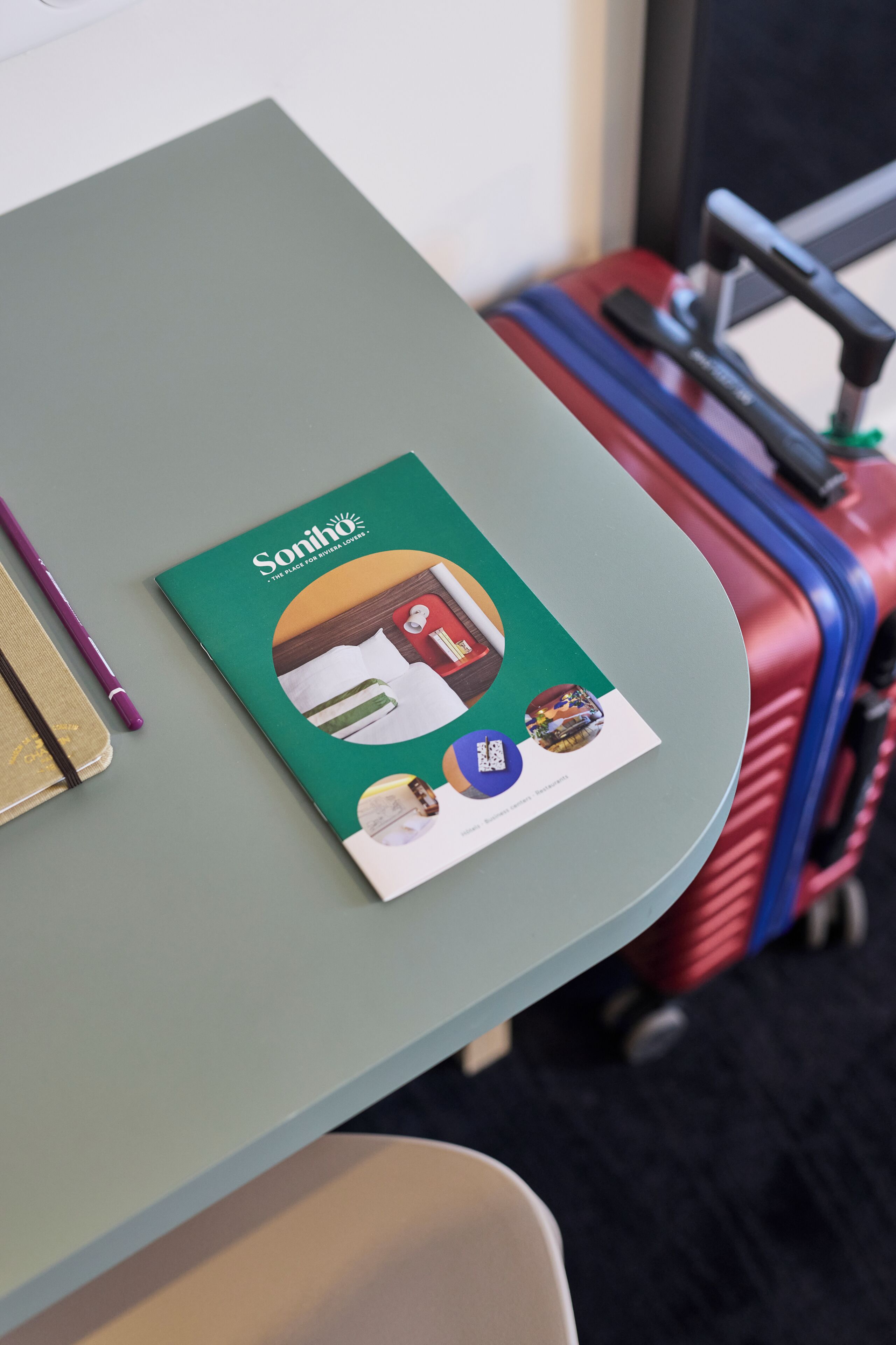 A green table with a brochure, a purple pen, and a red suitcase.