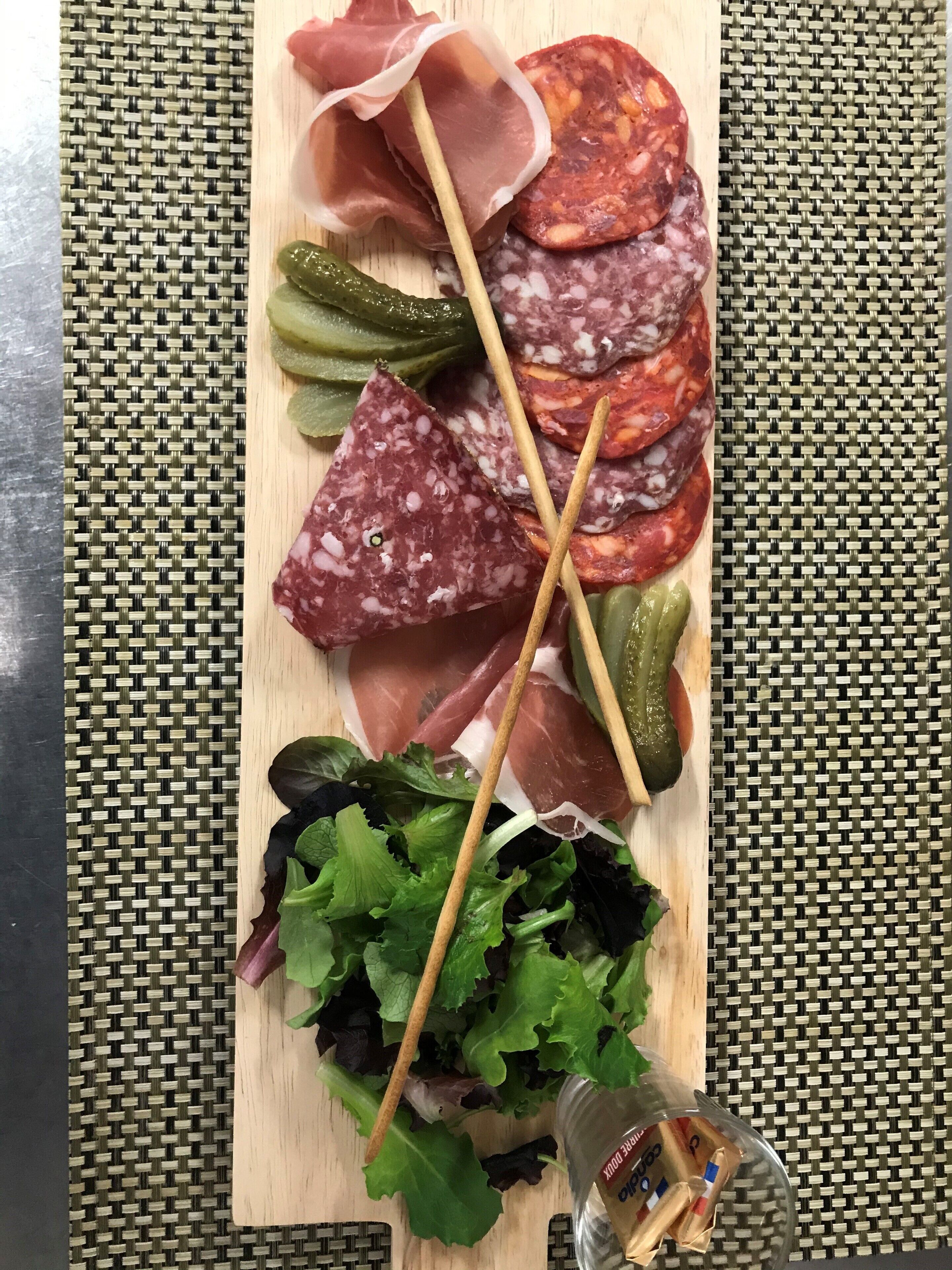 A wooden board with various cured meats, pickles, and leafy greens.