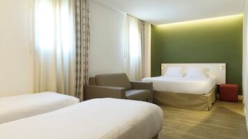 Camera d'albergo con due letti, una poltrona, un poggiapiedi rosso e una parete verde decorativa.