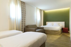 Camera d'albergo con due letti, una poltrona, un poggiapiedi rosso e una parete verde decorativa.