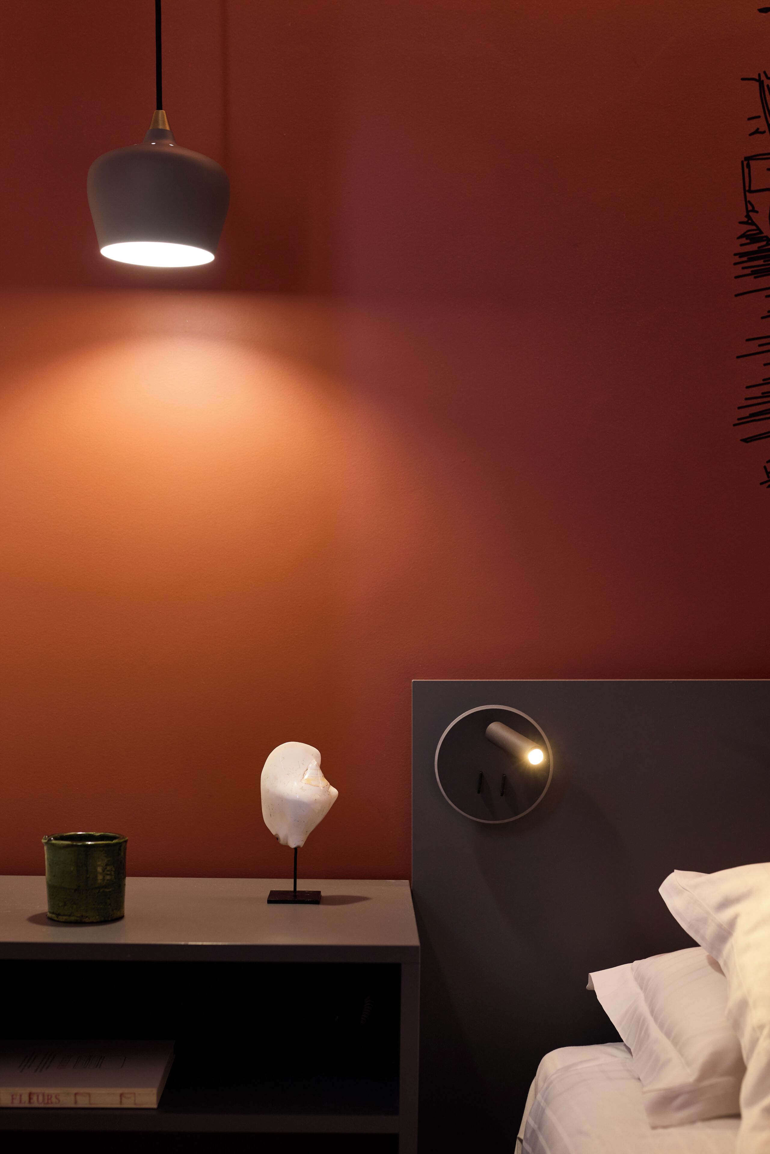 A room with a bed, a bedside table with a lamp, a wall clock, and a shelf with a book.