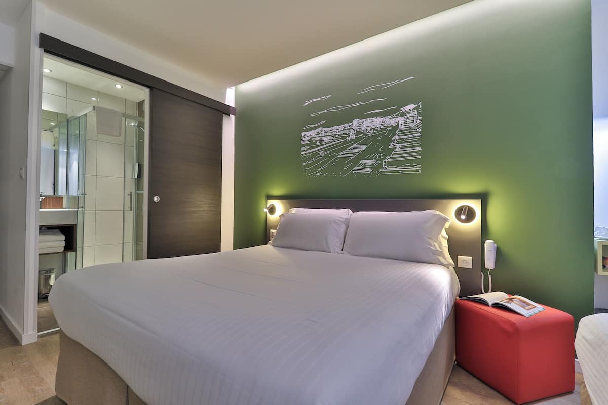 A modern bedroom with a large bed, a red ottoman, and a wall-mounted light fixture.