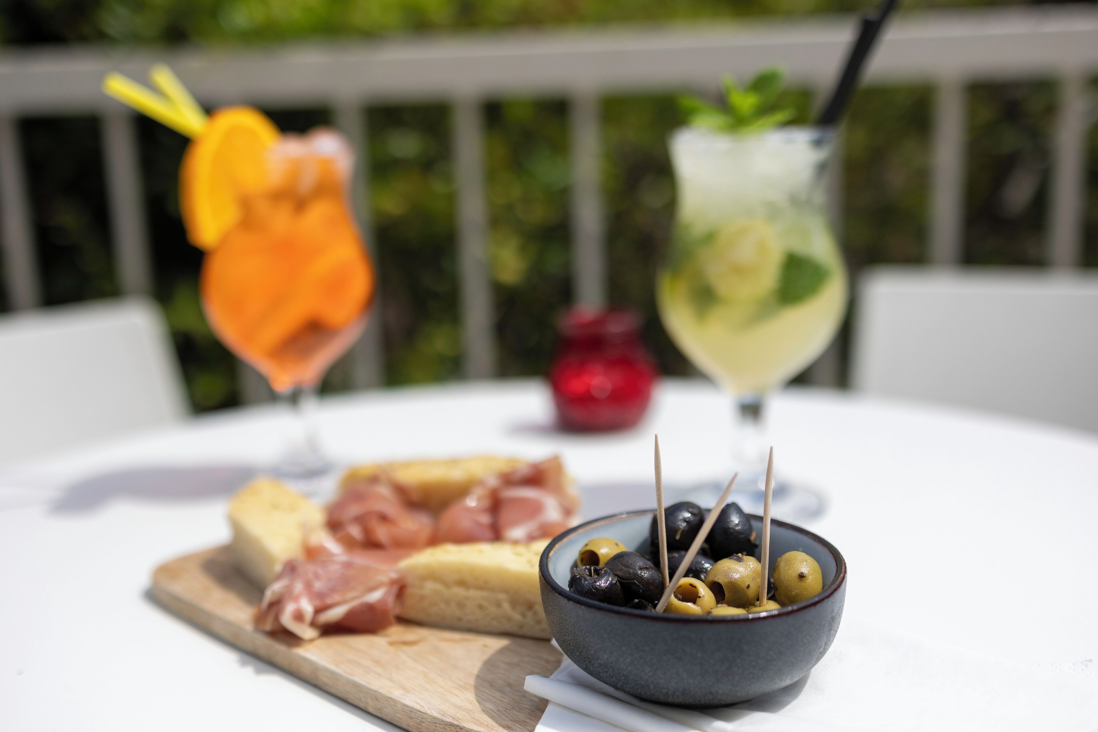 A table with a bowl of olives, a glass of drink with a straw, and a wooden board with cheese and meat.