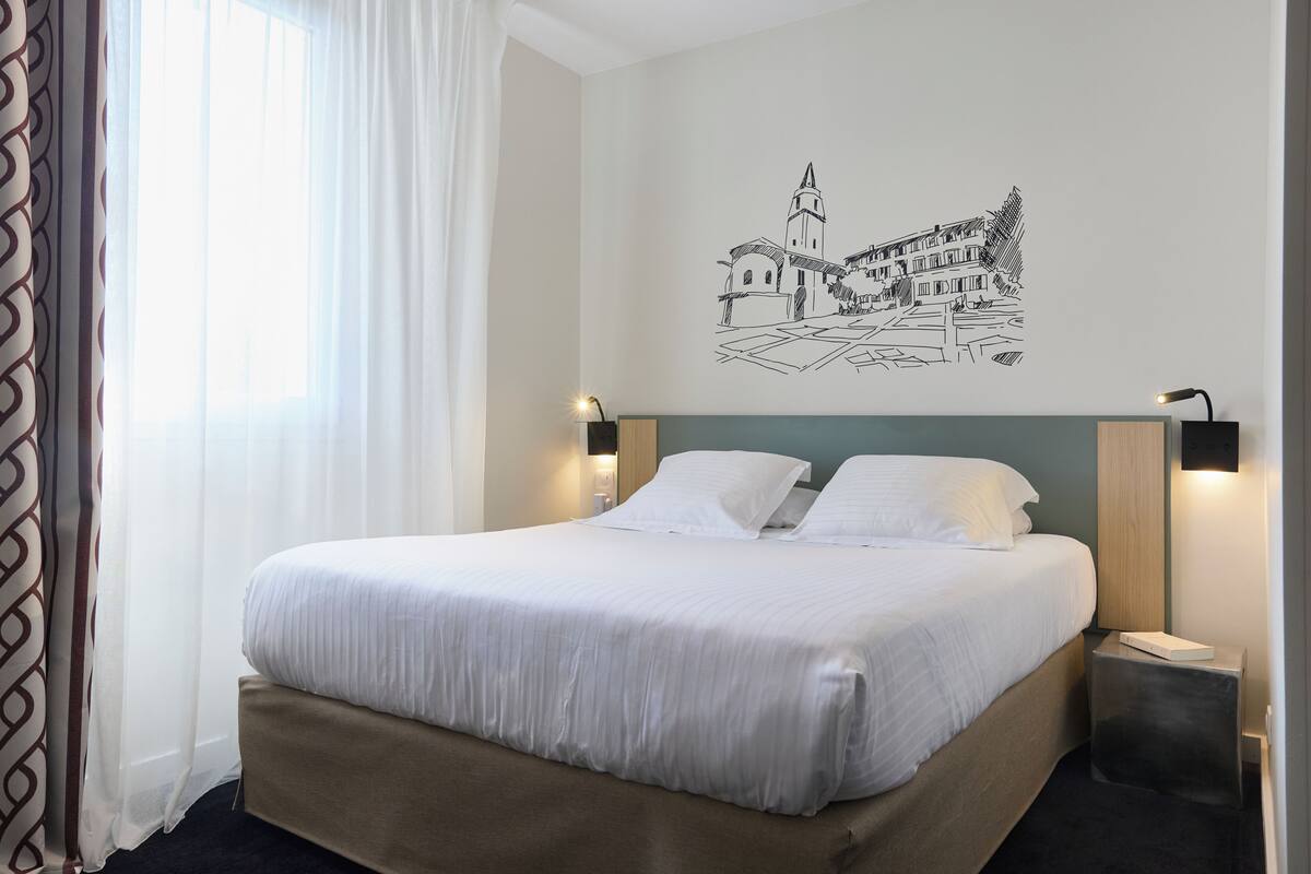 A hotel room with a bed, a wall-mounted artwork of a town, and two bedside lamps.