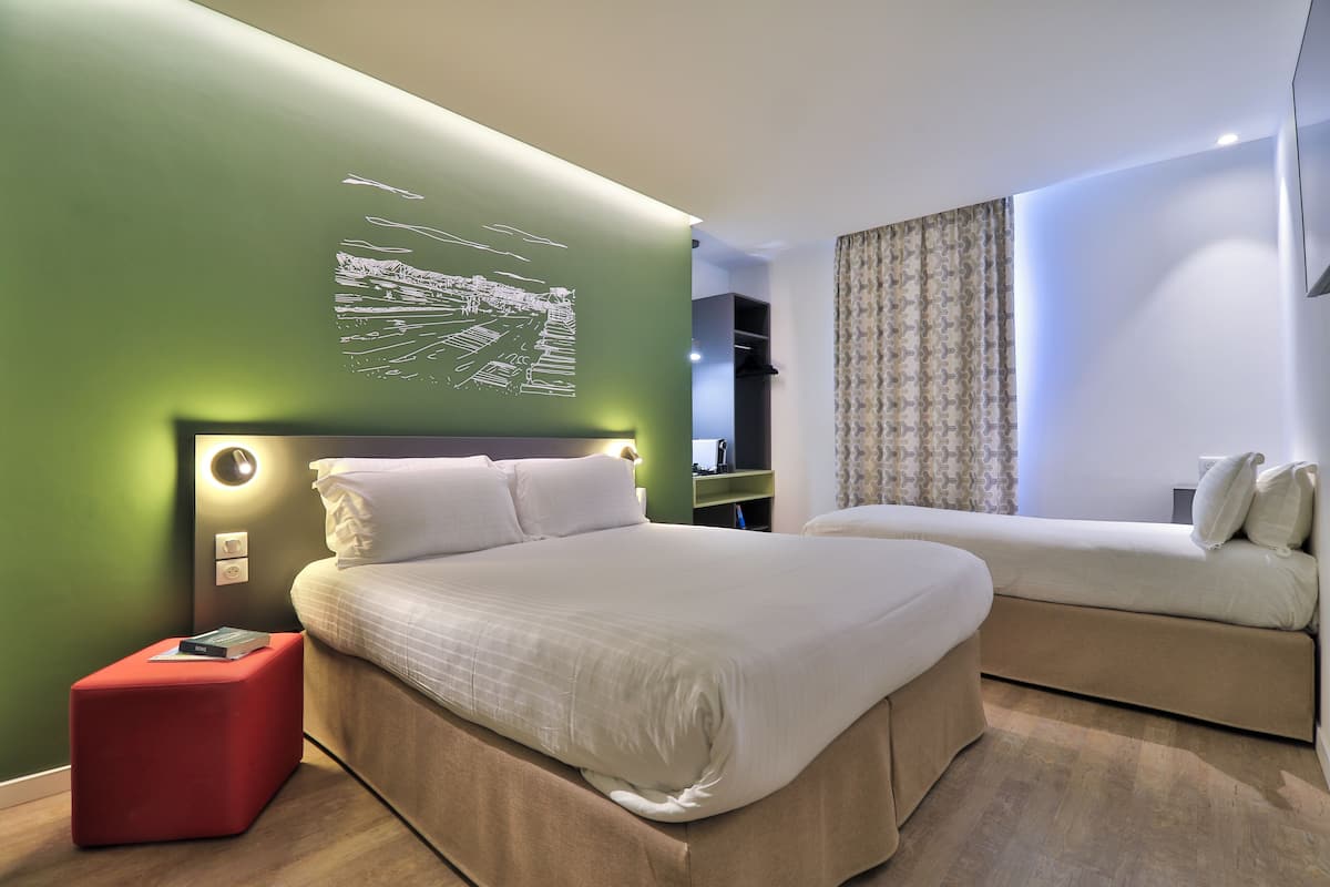 A modern hotel room with a large bed, a smaller sofa, a red ottoman, and a wall-mounted artwork.