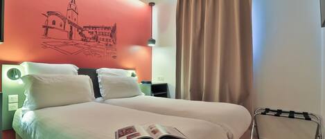 Quarto de hotel com uma cama grande, duas almofadas, mesa de cabeceira com abajur, parede com um mural vermelho e janela com cortinas.