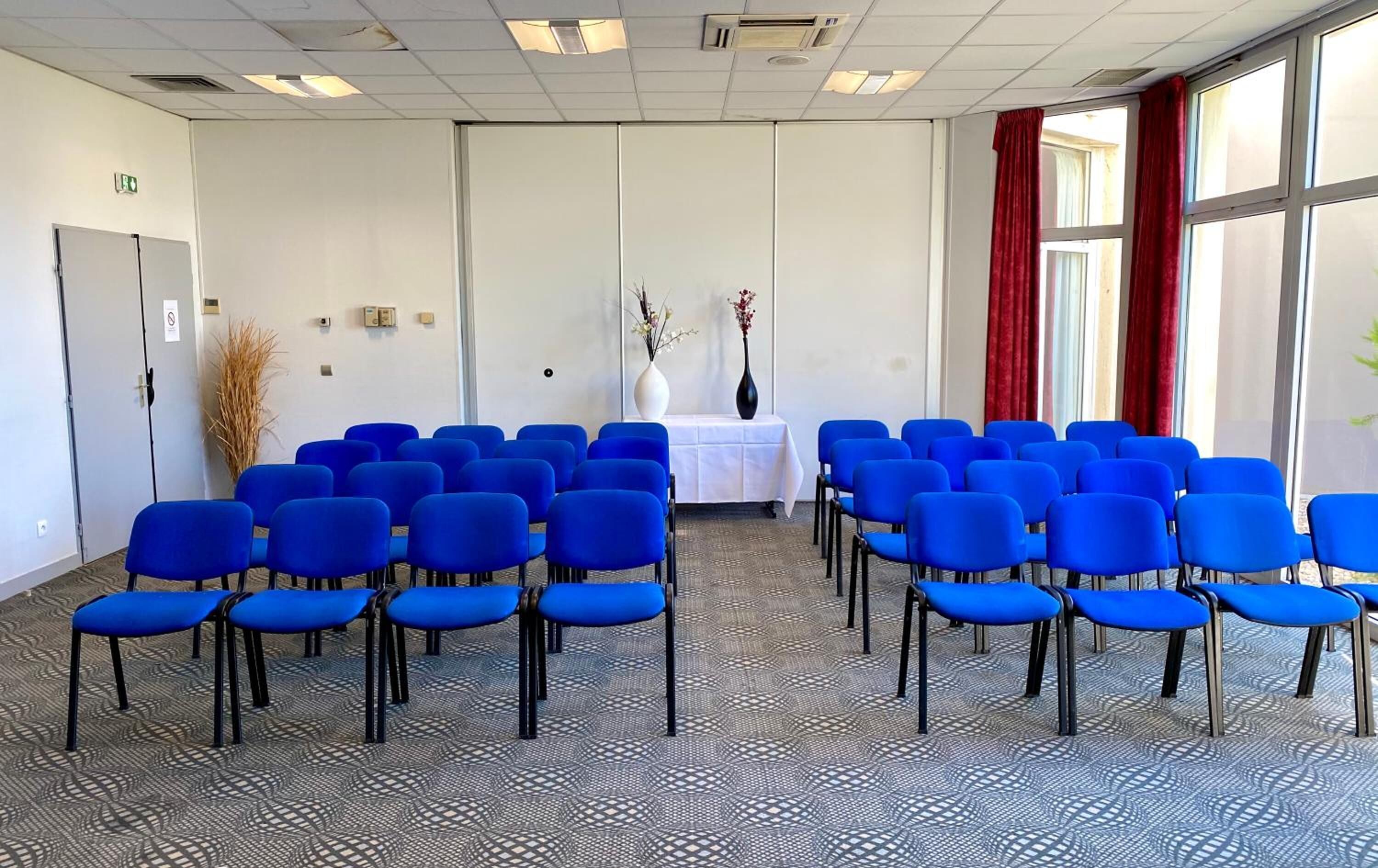 Un salón de conferencias con filas de sillas azules, una mesa blanca y un florero con flores.