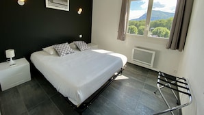 A bedroom with a bed, nightstand, wall-mounted light, window with curtains, and a view of greenery and mountains. - Hôtel le Néron (Fontanil-Cornillon)
