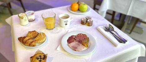 Une table de petit-déjeuner avec des croissants, un muffin, une assiette de jambon et de salami, un verre de jus d’orange, une tasse de café et divers condiments.