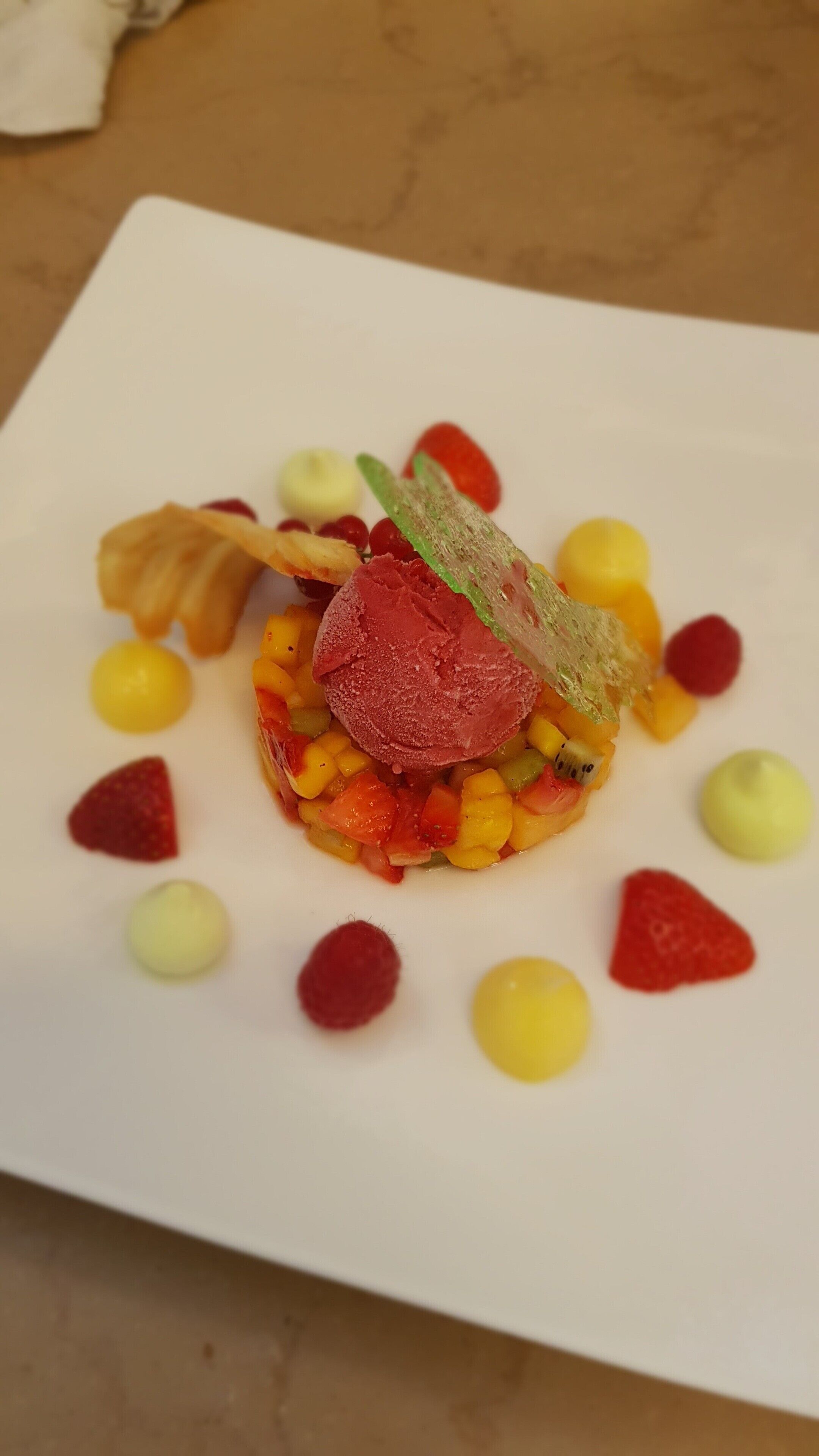 A square white plate with a colorful fruit and vegetable salad, garnished with small dollops of yellow and red sauces.
